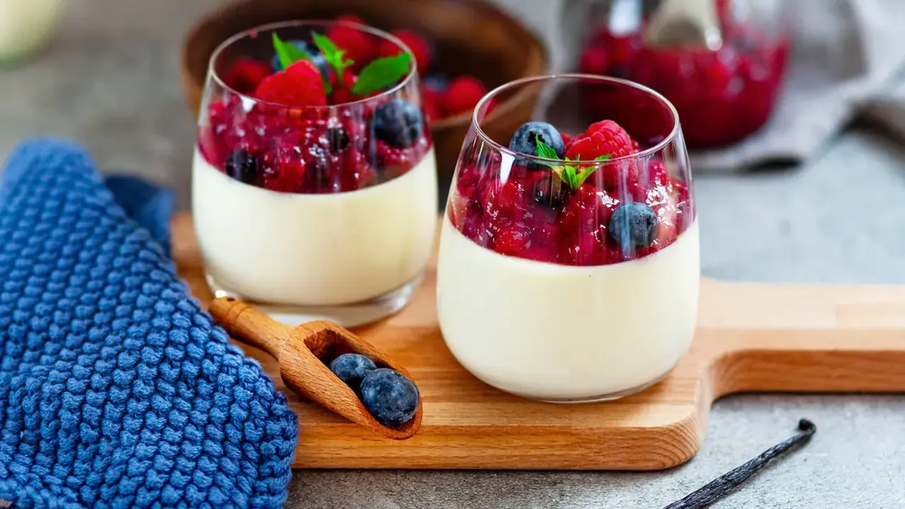 Zwei Gläser cremiger Panna Cotta mit Beerenkompott und frischen Beeren, garniert mit Minze. Ein Holzlöffel mit Blaubeeren liegt daneben.