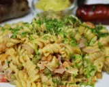 Ein cremiger nudelsalat mit mayo, garniert mit Schnittlauch. Daneben liegen zwei gebratene Würstchen und ein Schälchen Senf.