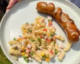 Gegrillte Wurst mit einem cremigen Nudelsalat als Beilage, garniert mit Erbsen, Mais und gewürfeltem Schinken.