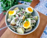 Schüssel mit cremigem Kartoffelsalat mit Gurken, Zwiebeln und gekochten Eiern, garniert mit frischen Kräutern.