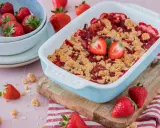 Ein frisch gebackener Erdbeer Crumble in einer hellblauen Auflaufform, garniert mit Erdbeerhälften. Daneben eine Schüssel voller Erdbeeren.