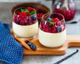 Zwei Gläser cremiger Panna Cotta mit Beerenkompott und frischen Beeren, garniert mit Minze. Ein Holzlöffel mit Blaubeeren liegt daneben.