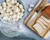 Würfel und Scheiben von Tofu liegen neben einem Messer auf einem Holzbrett. Die Frage "Kann man Tofu roh essen?" wird gestellt.