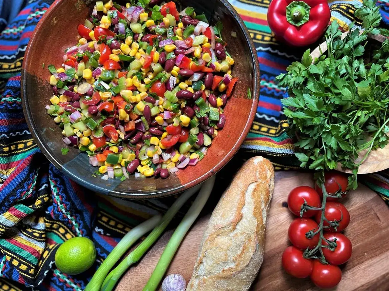 Bunter Salat zum Grillen mit Mais, Bohnen, Tomaten und Gurken, daneben Baguette, Kräuter und Gemüse.