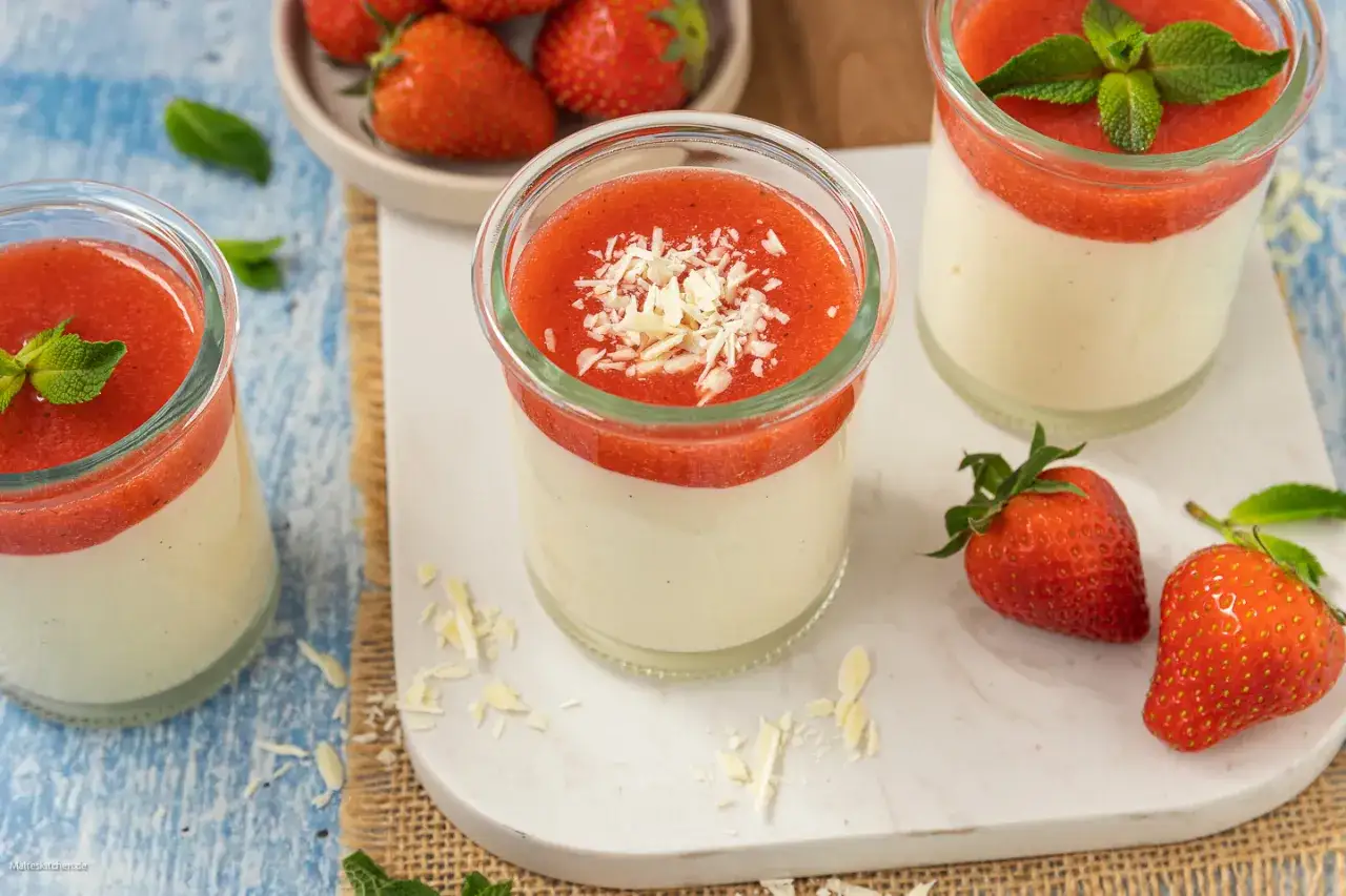 Cremige Panna Cotta mit Erdbeersoße und weißen Schokostreuseln, garniert mit Minze. Frische Erdbeeren liegen daneben.