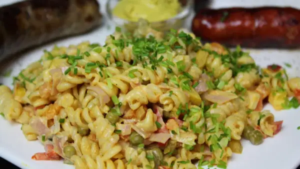 Ein cremiger nudelsalat mit mayo, garniert mit Schnittlauch. Daneben liegen zwei gebratene Würstchen und ein Schälchen Senf.