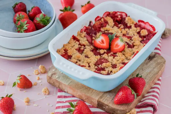 Ein frisch gebackener Erdbeer Crumble in einer hellblauen Auflaufform, garniert mit Erdbeerhälften. Daneben eine Schüssel voller Erdbeeren.