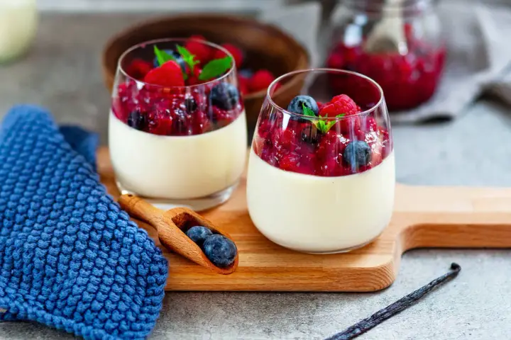 Zwei Gläser cremiger Panna Cotta mit Beerenkompott und frischen Beeren, garniert mit Minze. Ein Holzlöffel mit Blaubeeren liegt daneben.